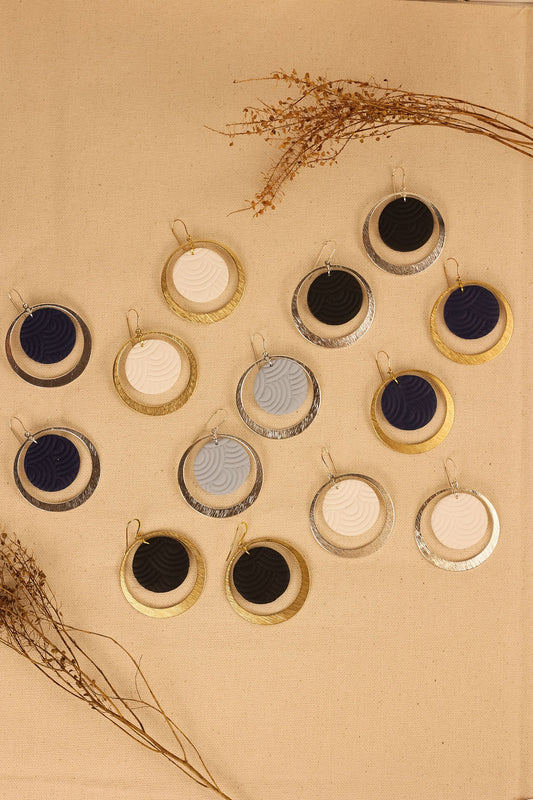 A collection of circular clay earrings in various colors including gold, silver, black, and white, displayed on a beige background.