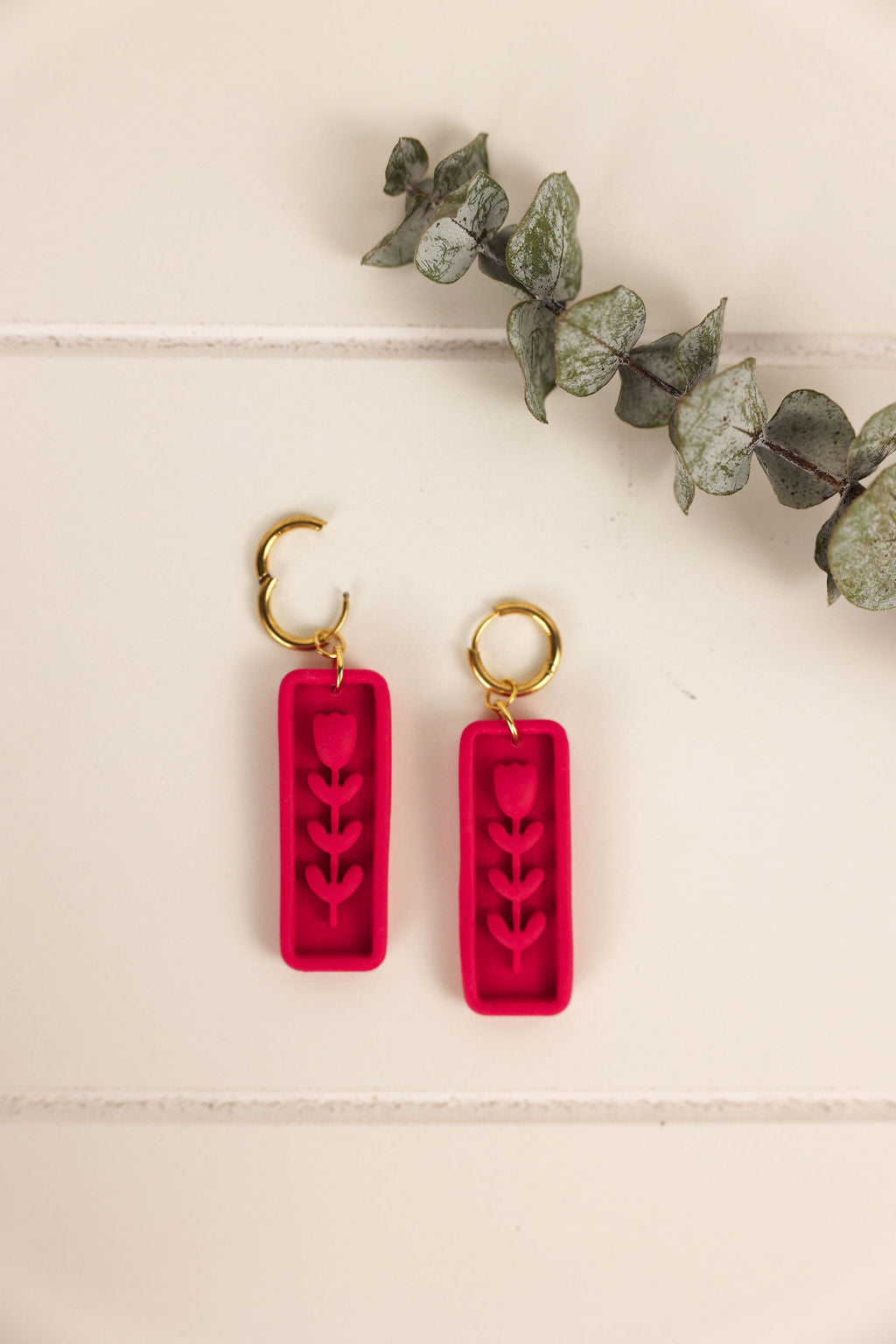 Hot pink earrings with gold hooks on a light wooden surface with eucalyptus leaves.