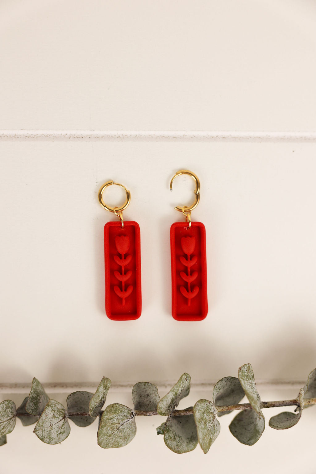Red earrings with gold hooks on a white background with green leaves at the bottom.