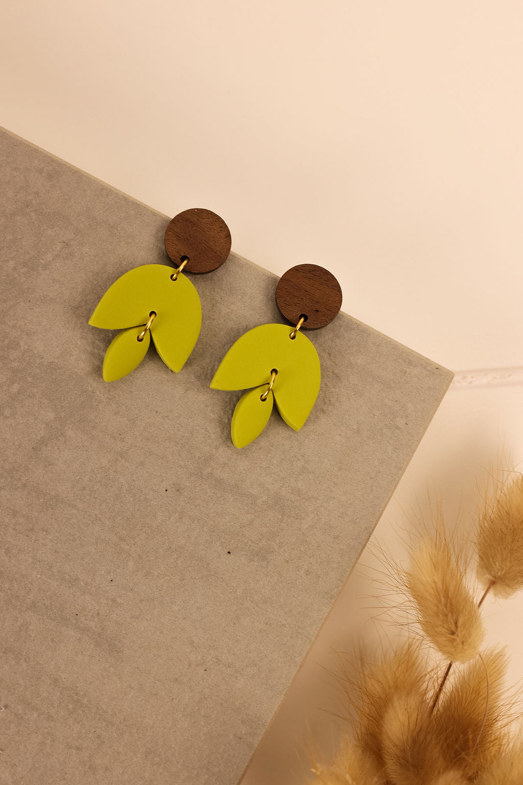 A pair of green clay earrings with a tulip design, displayed on a grey stand, with a beige object in the foreground.