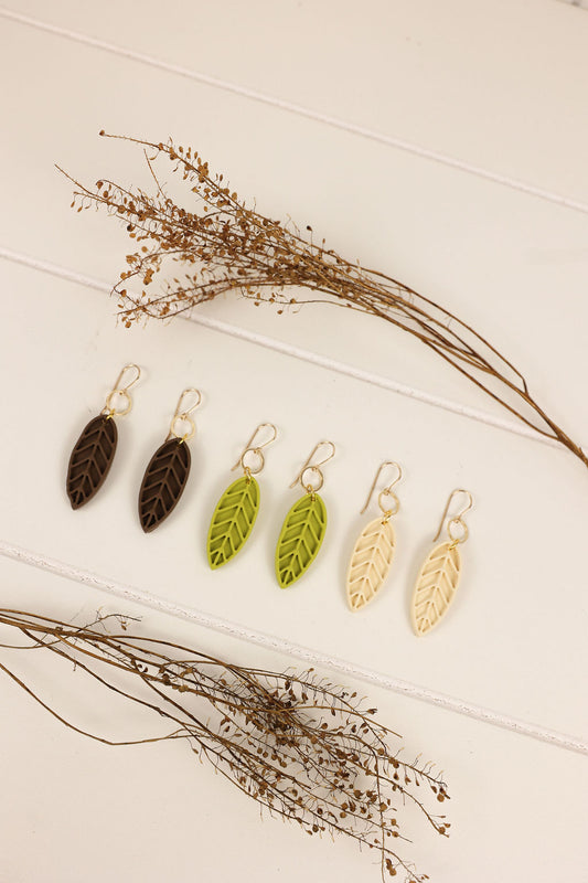 Set of leaf-shaped earrings on a wooden surface with dried plants.