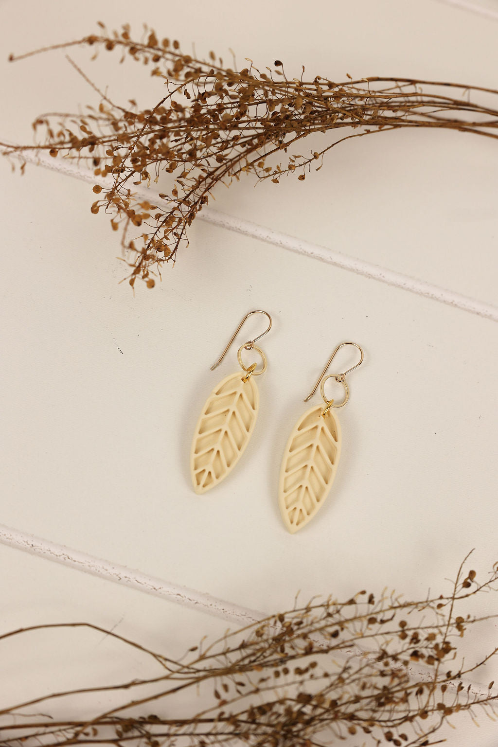 Ivory colored leaf earrings on a light background with dried branches.
