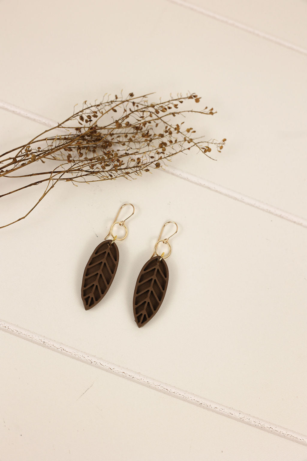 Pair of leaf-shaped earrings on a light surface with dried plants.