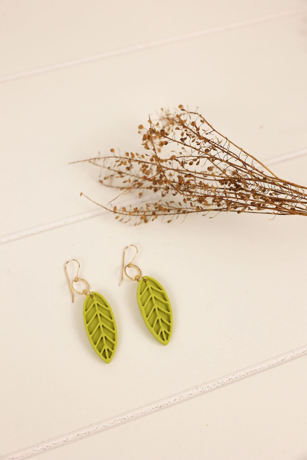Green leaf-shaped earrings on a beige surface with dried flowers.