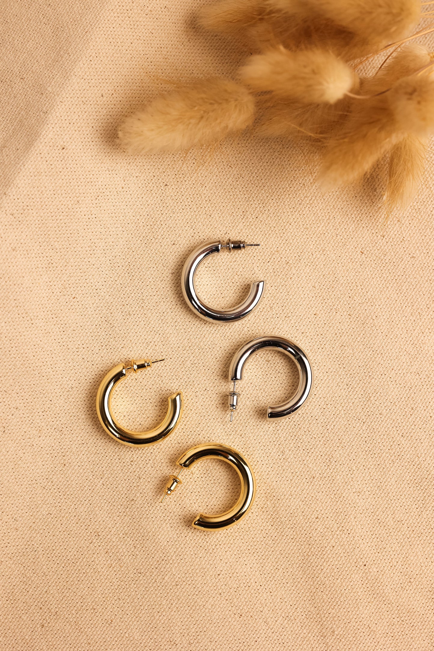 A collection of hollow hoop earrings in gold and silver colors, displayed on a textured surface next to a decorative feather.