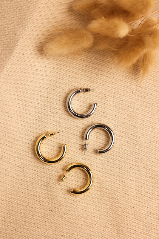 A collection of hollow hoop earrings in gold and silver colors, displayed on a textured surface next to a decorative feather.