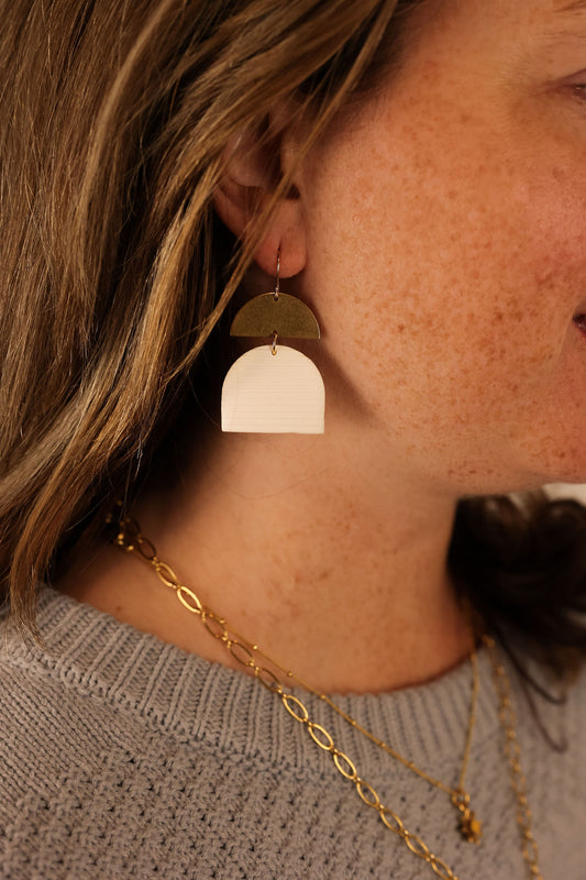 A person wearing the Molly clay earrings, which are composed of beige hand-rolled clay, a brass half circle charm, and a gold-plated sterling silver ear wire.