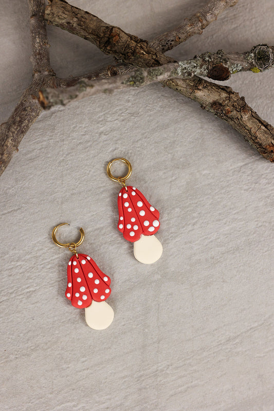Red and white polka dot mushroom earrings with gold hardware on a textured surface.
