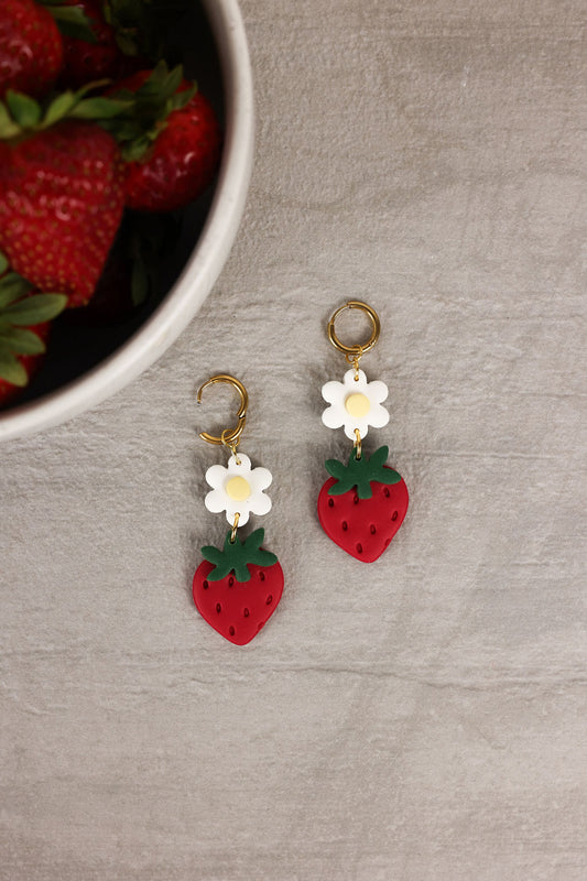 Strawberry and flower earrings on a gray surface with strawberries in the background.