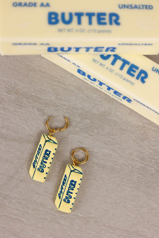 Butter container-shaped earrings on a surface with butter packages in the background