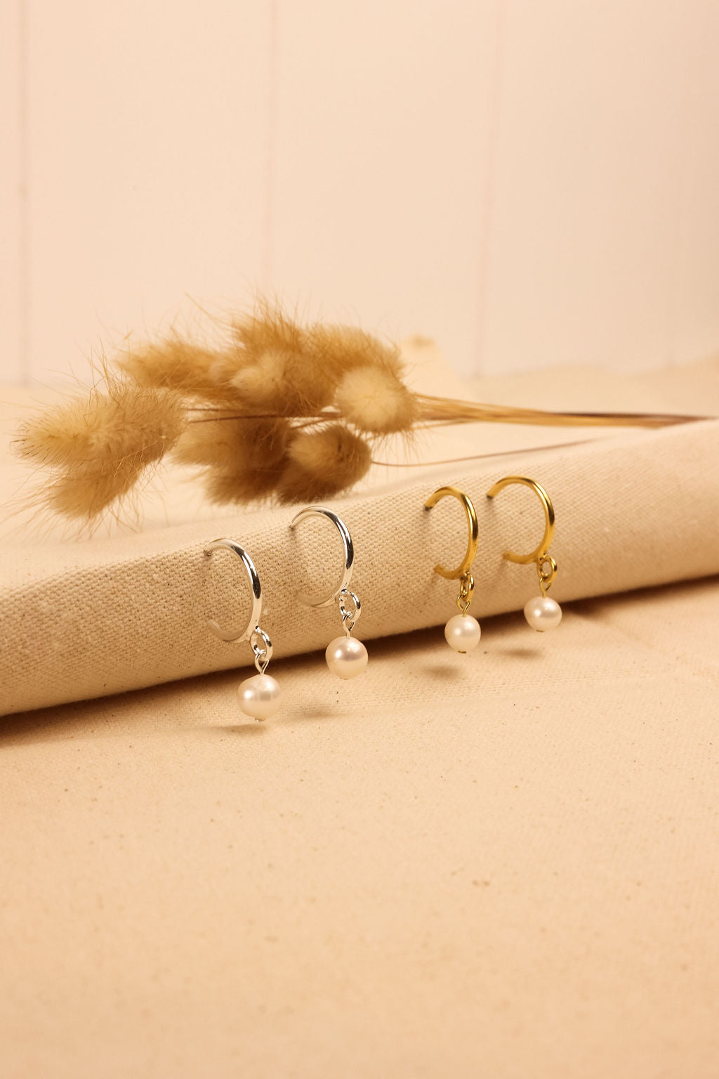 A pair of gold and silver colored pearl hoop earrings displayed on a textured surface, with a small feather and a book in the background.