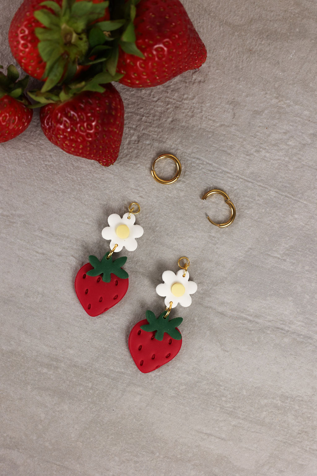 Strawberry-themed earrings with flowers on a gray surface with strawberries in the background.