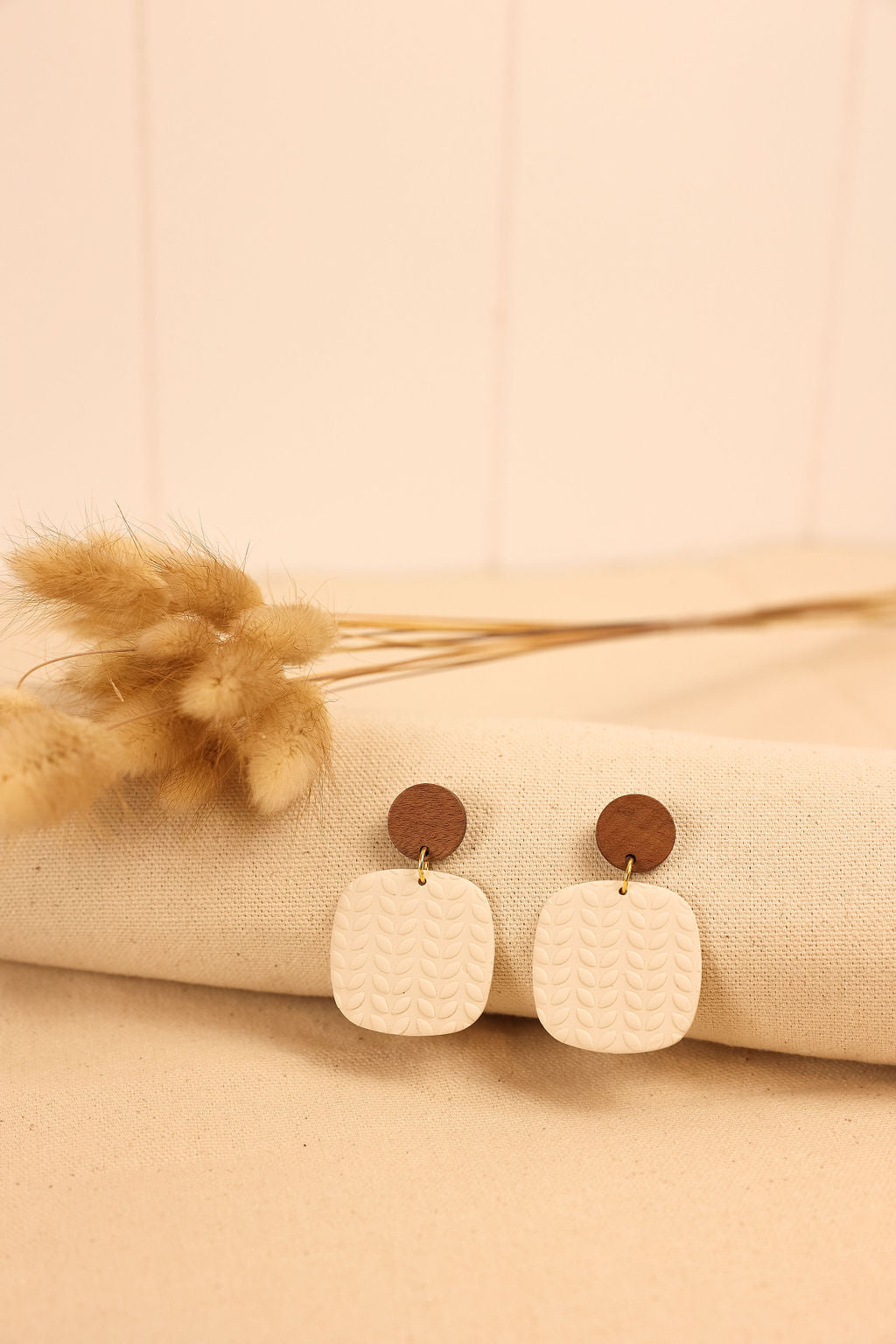 A pair of creamy white clay square earrings with a wood top, displayed on a textured surface.