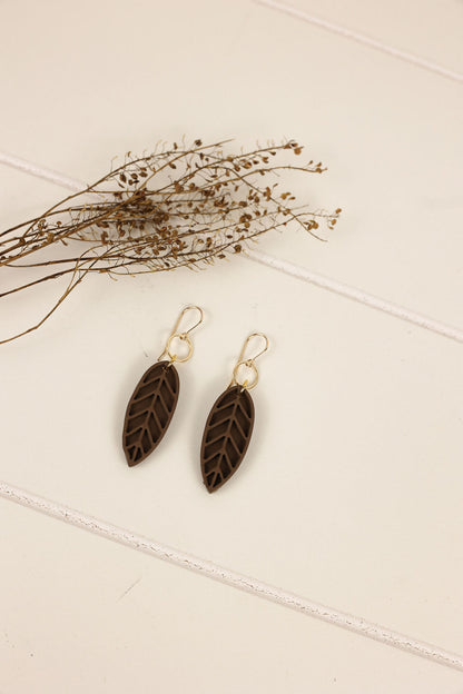 Pair of leaf-shaped earrings on a light surface with dried plants.