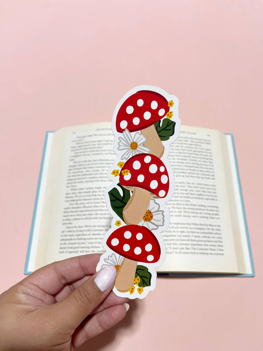 A die cut bookmark featuring a red and white mushroom design, held in front of an open book. 