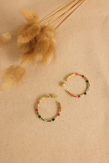 A pair of gold-colored hoop earrings with colorful enamel strips, displayed on a textured background.