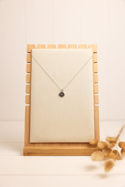 A crystal disc necklace with a tiny clear crystal in the center, displayed on a textured grey background, alongside a pair of dry reeds. The necklace is available in gold and silver colors.